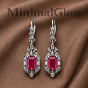 May include: A pair of silver-toned drop earrings featuring a central rectangular deep pink gemstone surrounded by small clear stones. The metalwork includes geometric patterns and the text MinimalGlow is visible at the top of the image.