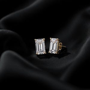 May include: A pair of gold stud earrings with rectangular, clear gemstones. The earrings are set against a black, silky background, creating a contrast that highlights the sparkle of the gems. The earrings are a classic jewelry item.
