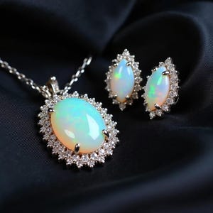 May include: A jewelry set featuring an opal pendant necklace and matching earrings. The oval opals have a milky, iridescent appearance, surrounded by a halo of small, sparkling diamonds. The jewelry is set against a dark, textured background.