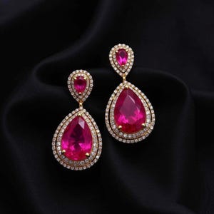 May include: A pair of gold-toned drop earrings with large, teardrop-shaped, fuchsia-colored gemstones. The gemstones are surrounded by a halo of small, clear crystals. The earrings are displayed on a black background.