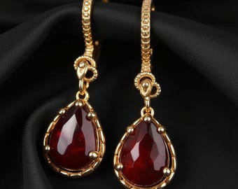 May include: A pair of gold-colored earrings with a teardrop-shaped, deep red gemstone. The earrings have a textured design on the hoops and around the gemstone. The gemstones are framed with a gold-colored border and small gold-colored beads.