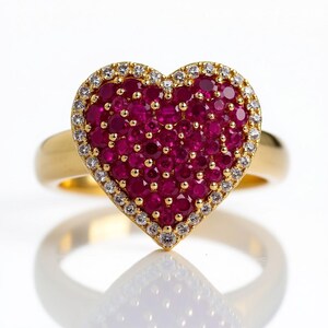 May include: A gold ring featuring a heart-shaped design. The heart is filled with numerous small, vibrant red gemstones, surrounded by a border of smaller, clear gemstones. The ring's band is a smooth, polished gold.