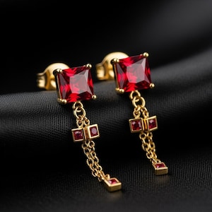 Peut inclure: Paire de boucles d'oreilles dorées avec des pierres rouges carrées. Chaque boucle d'oreille est composée d'une tige, d'une pierre rouge carrée, d'une chaîne et d'une pierre rouge carrée pendante. Sur fond noir.