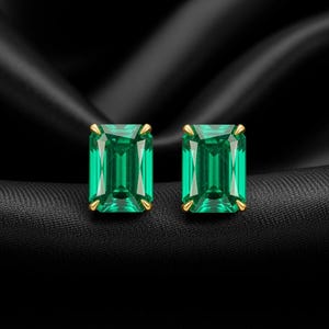 May include: A pair of emerald-cut, vibrant green gemstone stud earrings. Each earring is set in a gold-colored prong setting. The earrings are displayed on a black, textured surface, creating a striking contrast.