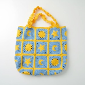 May include: A handmade crochet tote bag in a celestial pattern. The bag features a blue background with yellow squares, each containing a sun, moon, or star design. The handles are a matching yellow. The bag is designed for carrying items.