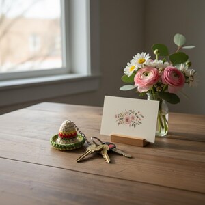 May include: A wooden table displays a small bouquet of pink and white flowers in a glass vase, a greeting card with a floral design, and a crochet sombrero keychain next to a set of keys.