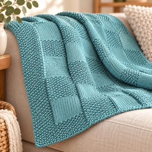 Könnte beinhalten: Eine blaugrüne Strickdecke mit einem strukturierten Schachbrettmuster aus abwechselnden glatten glatt rechts gestrickten Quadraten und körnigen kraus rechts gestrickten Quadraten, drapiert über einem neutralen Sofakissen.