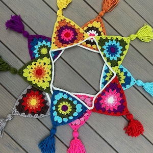 May include: A colorful, handmade crochet garland featuring triangular panels in various vibrant colors, including pink, yellow, blue, and green. Each panel has a central floral design and is adorned with a tassel at each point. The garland is displayed on a wooden surface.