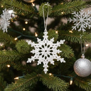 Peut inclure: Une décoration de Noël en forme de flocon de neige blanc crocheté est suspendue à un sapin de Noël vert. Le flocon de neige présente un motif ajouré délicat. D'autres décorations argentées et des lumières sont visibles en arrière-plan.