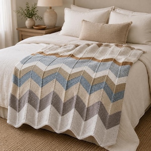 May include: A knitted throw blanket featuring a repeating chevron pattern in shades of cream, light blue, tan, and grey. The blanket has a textured, ribbed knit construction with vertical lines separating the chevron sections across the fabric.