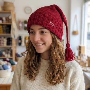 May include: A red knit hat with a long tassel and the word "Etsy" embroidered on the side. The hat is a deep red color and has a ribbed texture. The tassel is made of the same material as the hat.
