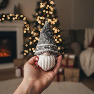 Op de afbeelding: Een handgemaakte gehaakte kabouter met een grijze hoed, witte baard en rood-wit gestreept lichaam. De kabouter wordt in een hand gehouden, met een kerstboom en een open haard op de achtergrond. Een feestelijke vakantieversiering.