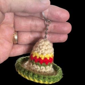 May include: A crocheted sombrero keychain in cream, green, yellow, and red. The miniature hat features a wide brim and a decorative band. The keychain is attached to a silver-colored ring and chain.