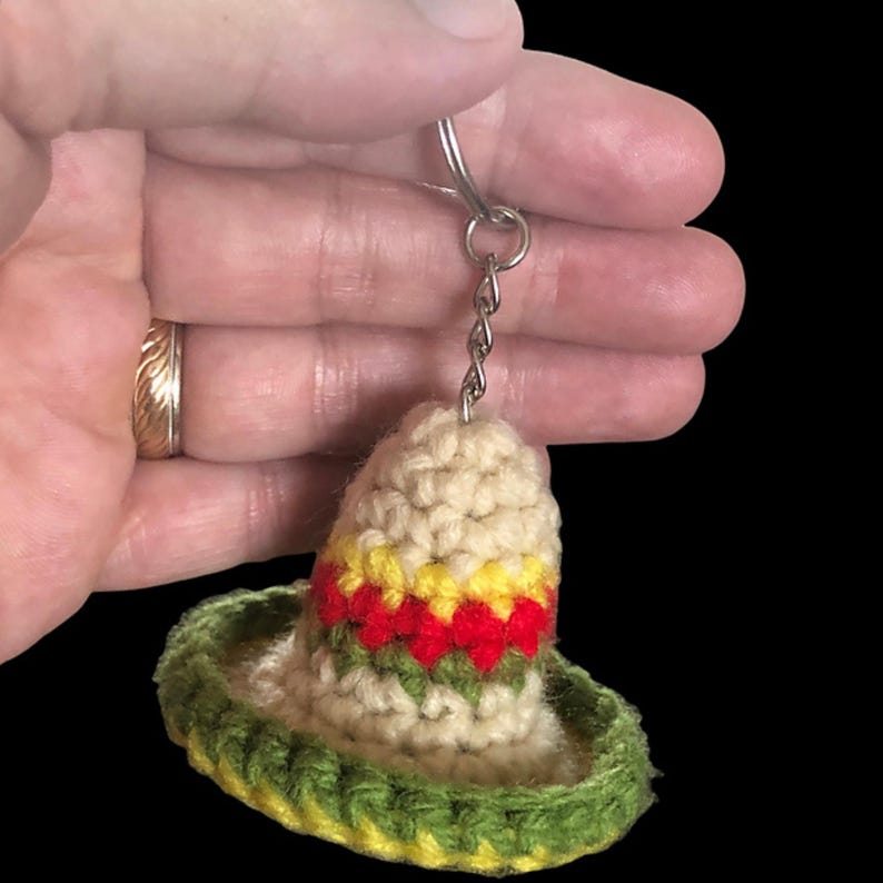 May include: A crocheted sombrero keychain in cream, green, yellow, and red. The miniature hat features a wide brim and a decorative band. The keychain is attached to a silver-colored ring and chain.