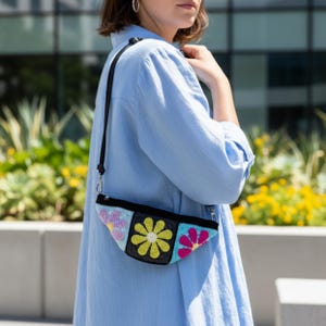 May include: A black crossbody bag with a colorful crochet floral design. The bag features three square panels with pink, yellow, and blue flowers. The strap is black and adjustable, and the bag is worn over the shoulder.