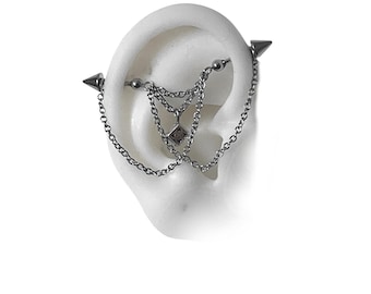 May include: A silver industrial barbell earring with chain and charm accents. The earring features a chain that drapes down from the barbell, with a small square charm. The barbell ends have cone-shaped spikes.