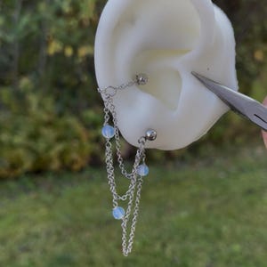 Peut inclure: Boucle d'oreille en chaîne argentée avec trois brins, chacun orné d'une petite perle bleu clair. La boucle d'oreille est fixée à un clou et reliée à un autre clou par une chaîne. La boucle d'oreille est présentée sur un modèle d'oreille blanc.