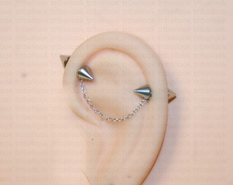 Op de afbeelding: Een zilveren oorring met een ketting die twee puntige uiteinden verbindt. De oorring wordt gedragen op de bovenste oorlel.