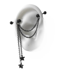 May include: Black industrial barbell earring with dangling chains and star charms. The earring has a straight barbell design with two ball ends, connected by chains that hang down, each ending with a star charm. The jewelry is shown on a white ear model.