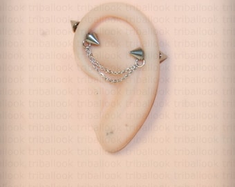 Op de afbeelding: Een zilveren oormanchet met twee spikes en een ketting die de spikes verbindt. De manchet wordt gedragen op het bovenste deel van het oor.