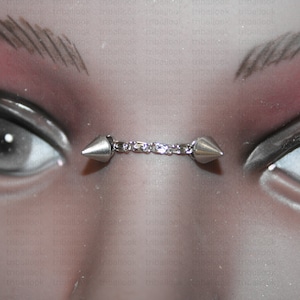 以下が含まれることがあります： 2つの尖った先端がチェーンでつながったシルバーの鼻ピアス。モデルに装着されています。