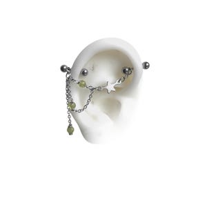 May include: Silver industrial barbell earring with a chain and star charm, adorned with small green beads. The earring is shown on a white ear model, showcasing the design and details.