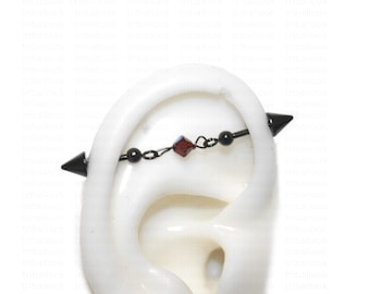 Peut inclure: Barbell industriel noir avec une perle de cristal rouge et des pointes noires à chaque extrémité. Le barbell est porté dans une fausse oreille.