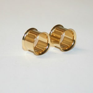 May include: Two gold-coloured metal ear gauges, one on the left and one on the right, are shown on a white surface. The gauges are round and have a smooth, polished finish.