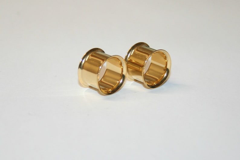 May include: Two gold-coloured metal ear gauges, one on the left and one on the right, are shown on a white surface. The gauges are round and have a smooth, polished finish.