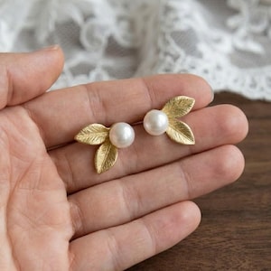 Puede incluir: Un par de pendientes dorados con forma de hoja, cada uno con una perla blanca en el centro. Los pendientes se muestran en una mano, mostrando su delicado diseño y estilo elegante. Las hojas tienen vetas detalladas.
