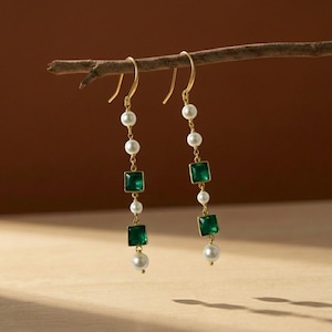 May include: A pair of gold-toned drop earrings. Each earring features a series of white pearls and square, emerald-green gemstones. The earrings are suspended from a curved hook and are displayed against a neutral background.