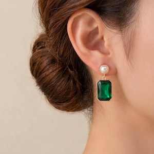 May include: A gold-toned earring featuring a small, round white pearl stud at the top, connected to a large, rectangular, emerald-green faceted gemstone pendant. The earring is suspended from a gold metal setting that holds the green stone securely.