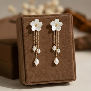 Puede incluir: Un par de pendientes elegantes con flores blancas y detalles dorados. Cadenas doradas cuelgan tres perlas blancas en forma de lágrima. Los pendientes se exhiben en una caja de joyería marrón.