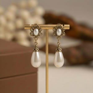 Puede incluir: Un par de pendientes de estilo vintage exhibidos en un soporte dorado. Cada pendiente presenta un centro de perla ovalado, detalles de filigrana ornamentados y una gota de perla en forma de lágrima. Diseño clásico y elegante.