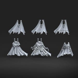 May include: A collection of six miniature grey plastic capes and cloaks for gaming figures. The items feature various designs including tattered edges, integrated mechanical cables, and decorative skull motifs, all rendered in a neutral matte grey finish.