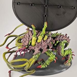 Könnte beinhalten: Detaillierte Miniaturfigur einer Kreatur mit grünem und braunem Exoskelett, roten Krallen und mehreren Tentakeln. Die Figur ist auf einem schwarzen Sockel montiert, mit einem schwarzen scheibenförmigen Element darüber.