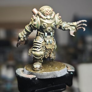Könnte beinhalten: Miniaturfigur eines Kriegers in Bronze- und grüner Rüstung. Die Figur hat einen Helm mit kreuzförmigem Visier und klauenartigen Händen. Die Rüstung ist mit Nieten, Ketten und geschichteten Platten detailliert. Die Figur steht auf einem kleinen, runden Sockel.