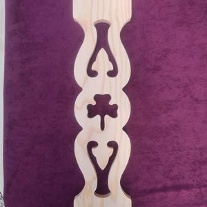 May include: A light-colored, unfinished wooden baluster with decorative cut-out designs. The baluster features a series of curved shapes and a central cloverleaf design. The wood grain is visible, and the piece is ready for staining or painting.
