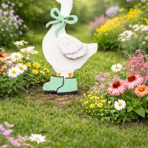 May include: A white wooden goose garden decoration with a green bow and boots. The goose has an orange beak and is standing in a garden with colorful flowers and green grass. A charming outdoor accent.