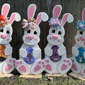 May include: Four wooden Easter bunny decorations. Each bunny is white with pink ears, nose, and paw pads. They hold decorated eggs and wear bows. The eggs have different designs, including crosses and floral patterns. They are displayed on wooden bases.
