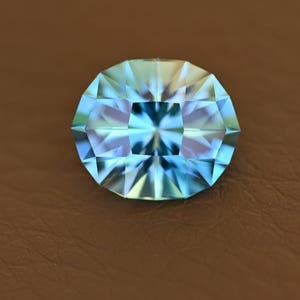 Op de afbeelding: Een ovaalvormige, gefacetteerde edelsteen met blauwe en groene tinten. De complexe slijping van de edelsteen creëert een sprankelend effect. De steen ligt op een bruine achtergrond.