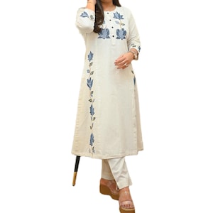 May include: Off-white, long-sleeved tunic and pants set. The tunic features blue floral embroidery along the front and sleeves. The pants are a matching off-white color. The outfit is a casual, yet elegant style.