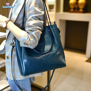 May include: A teal blue leather tote bag with a shoulder strap. The bag has a smooth, glossy finish and a simple, elegant design. The bag is being held by a person wearing a plaid blazer.