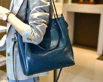 May include: A teal blue leather tote bag with a shoulder strap. The bag has a smooth, glossy finish and a simple, elegant design. The bag is being held by a person wearing a plaid blazer.