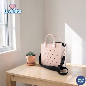 May include: A light pink tote bag with a black shoulder strap and handles. The bag has a perforated design and a zipper closure. A small potted succulent sits on a wooden table next to the bag. The brand name "Luvetote" is visible in the upper left corner.
