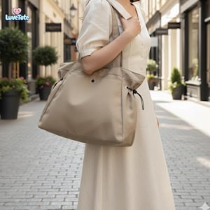 Peut inclure: Un sac fourre-tout beige avec une fermeture à cordon et un cordon de serrage noir. Le sac a un grand compartiment principal et une petite poche à l'avant. Le sac est porté par une personne vêtue d'une robe crème.