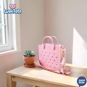 May include: A pink tote bag with a shoulder strap and handles sits on a wooden table. The bag has a perforated design and a zipper closure. A small potted succulent is next to the bag. The text "Luvetote" is in the upper left corner.