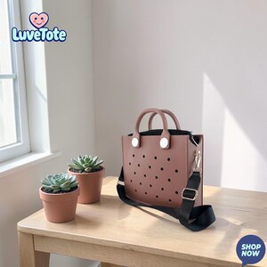 May include: A mauve tote bag with a black interior and a black shoulder strap. The bag has a series of circular perforations and two top handles. The logo "Luvetote" is in the upper left corner. Two small potted succulents are on the table.