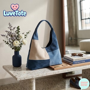 Puede incluir: Un bolso de hombro patchwork en tonos azules y crema, con una sola asa. El bolso se exhibe sobre una mesa de terrazo, junto a un jarrón con flores y una bandeja de madera con libros y gafas de sol. El logotipo de Luvetote es visible.