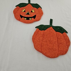May include: Two orange fabric pumpkin-shaped decorations with green leaf and stem accents. One pumpkin has a jack-o'-lantern face with black, yellow, and green details. The other pumpkin is plain.